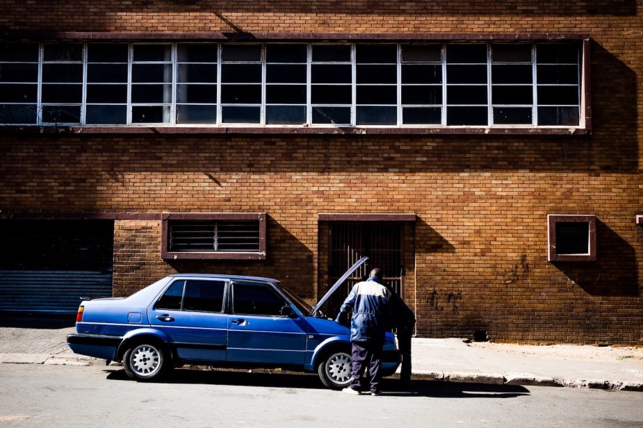 Two men look under the hood of an overheating car, a brick wall visible behind them.
