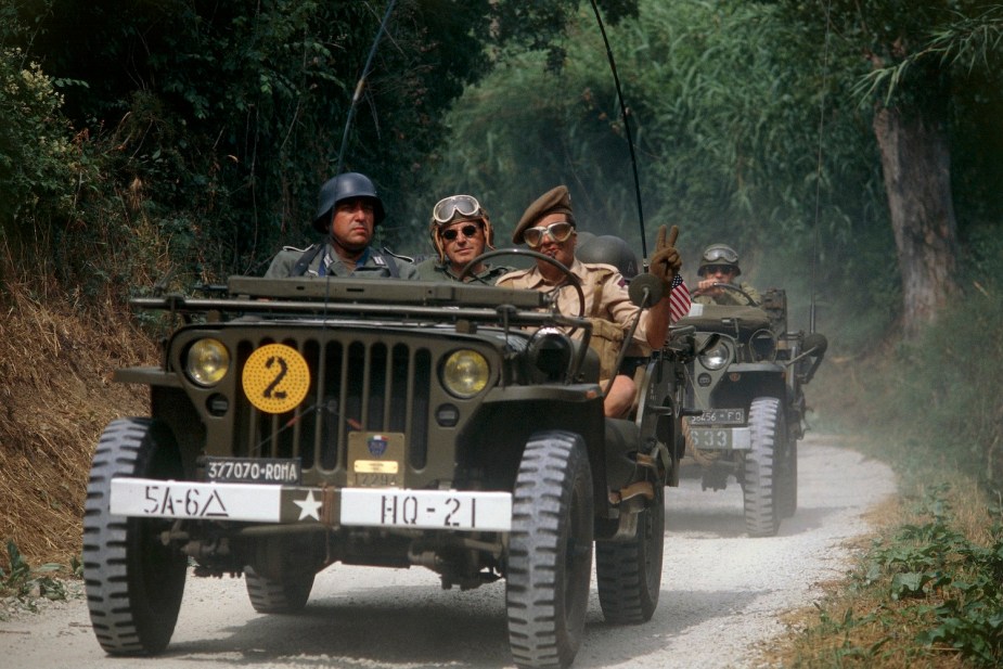 Reenactors drive down a dirt road in a 1941 Willys MB 4x4 Jeep, trees visible in the background.