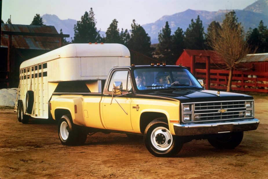 Full ton yellow and black square body Chevy truck towing a livestock trailer, a farm and mountain range visible in the background.