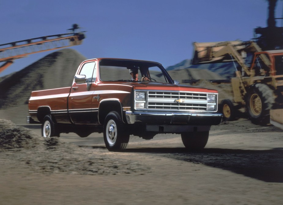 Promo photo of a 4x4 1987 square body Chevy truck, driving through a construction site with yellow equipment in the background.