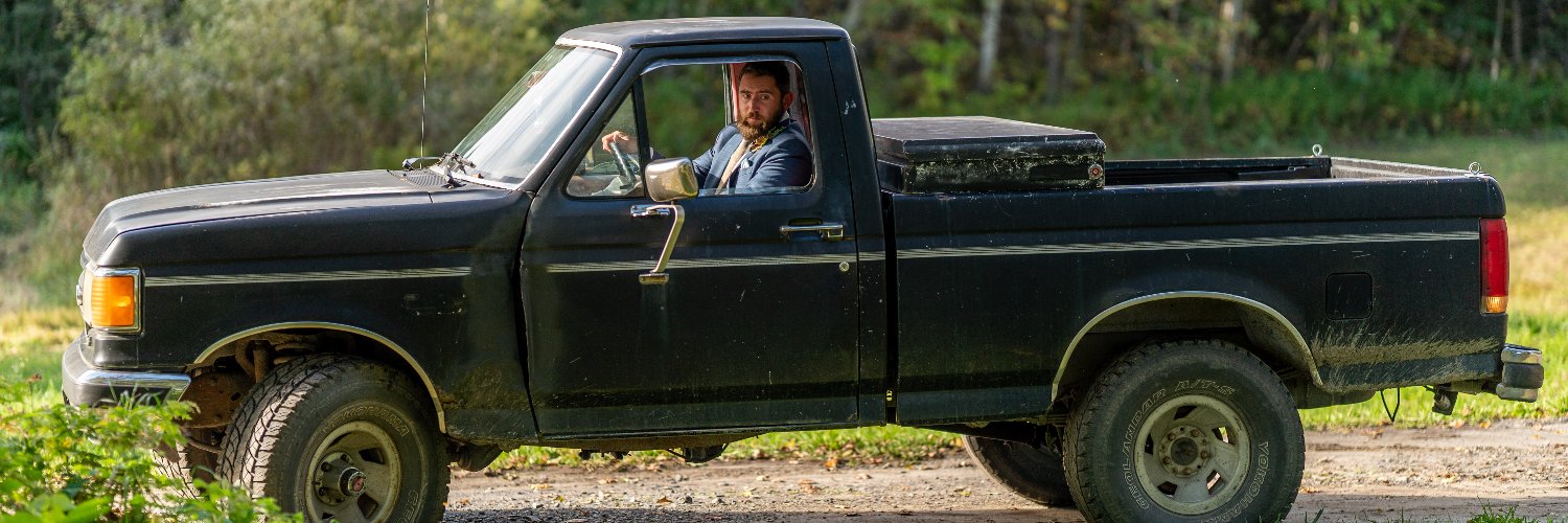 Automotive journalist Henry Cesari driving his classic square body Ford F-150 pickup truck, the woods of Vermont visible behind him.