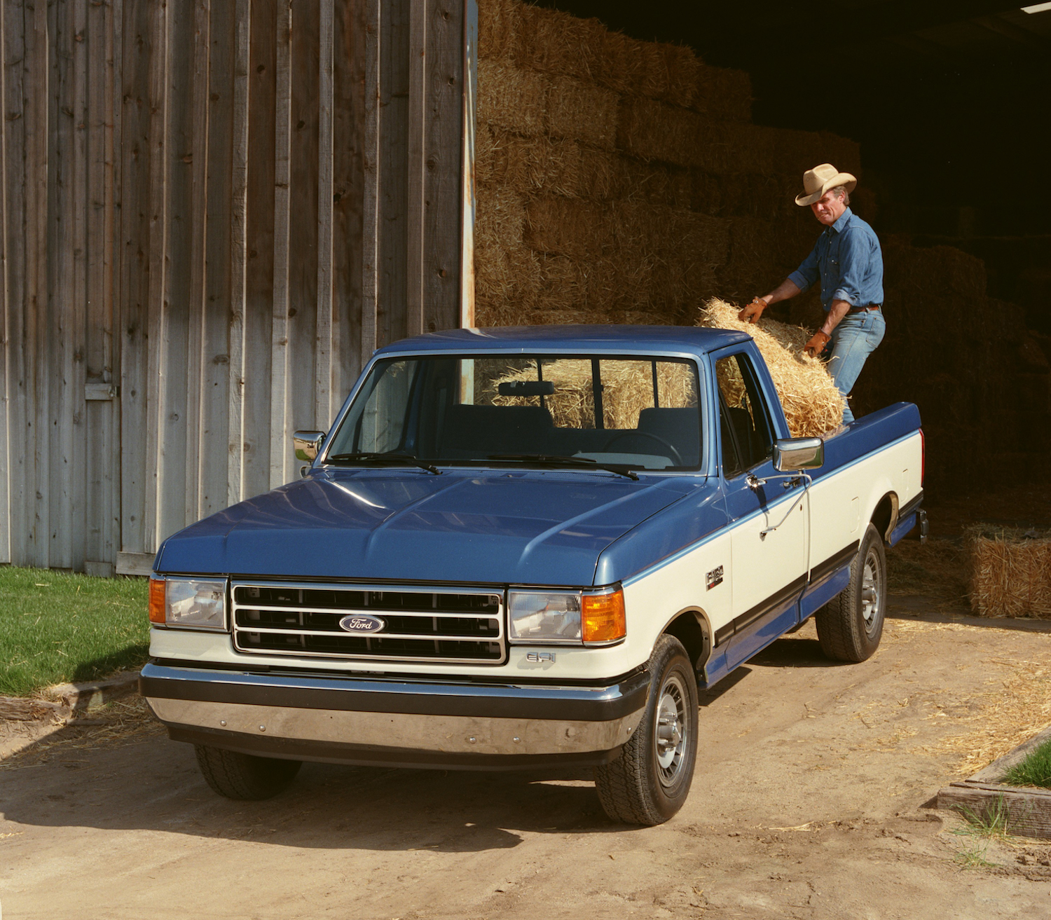 A two-tone blue and white square-body Ford pickup truck loaded with bales of hay, an open barn door visible in the background.