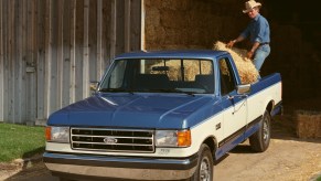 A two-tone blue and white square body Ford pickup truck being loaded with hay bales, an open barn door visible in the background.