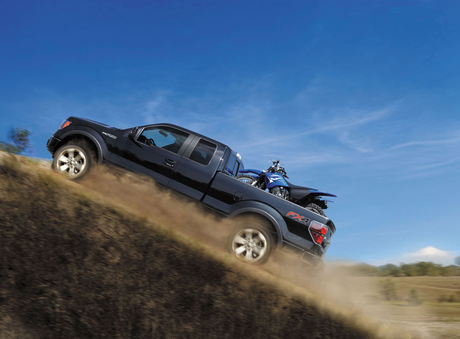 Promo photo of a 2014 Ford F-150 half-ton 4x4 pickup truck climbing a steep hill with a dirt bike in its bed, a blue sky visible in the background.