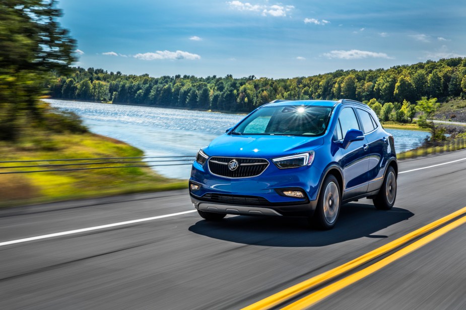 A bright blue Buick Encore crossover SUV driving past a lake and a forest on an American highway.