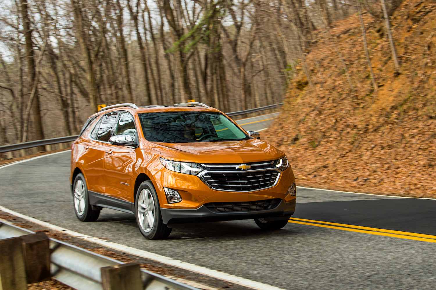 Orange Chevrolet Equinox crossover SUV driving around a curve in a rural road, trees and orange leaves visible in the background.