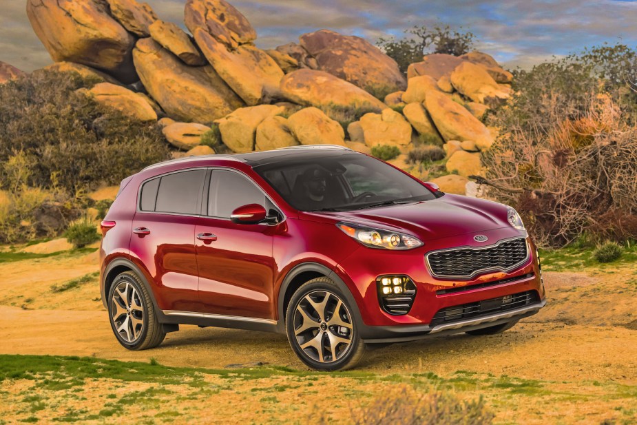 Red Kia Sportage crossover SUV parked on a dirt road, in front of a stack of tan-colored rocks and a cloudy sky.