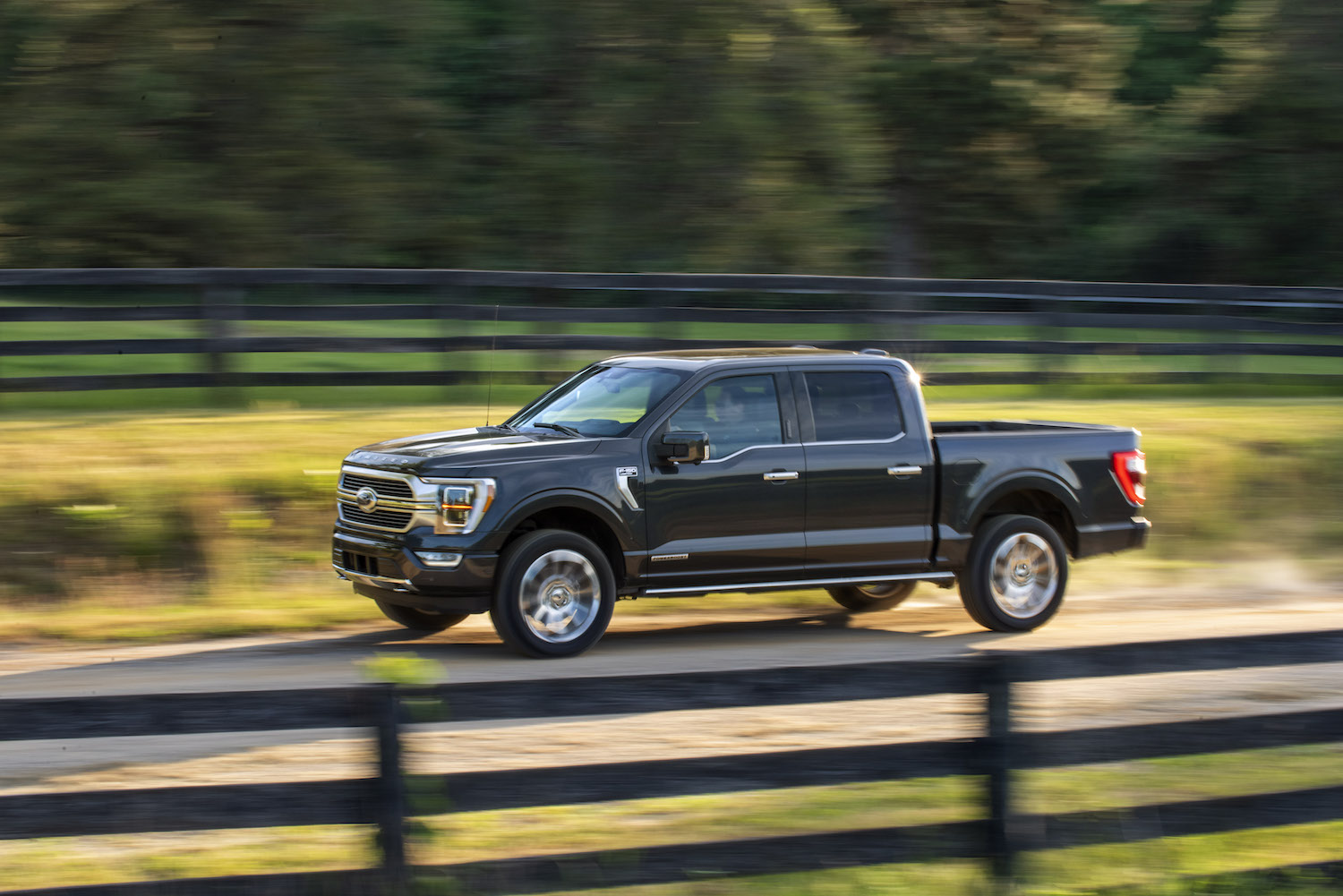 Promo photo of a dark gray Ford F-150 powerboost hybrid truck driving down a country road with a fence in the background.