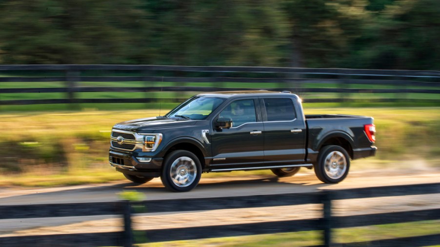 Promo photo of a dark gray Ford F-150 powerboost hybrid truck driving down a country road, a fence visible in the background.