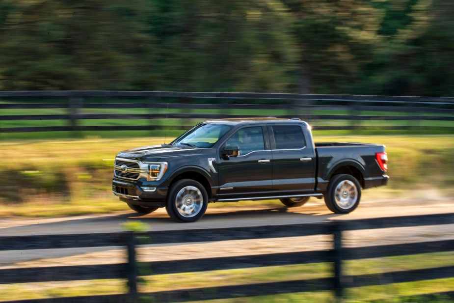 Promotional photo of a dark gray Ford F-150 Powerboost Hybrid Pickup truck driving on a country road, with a fence visible in the background.