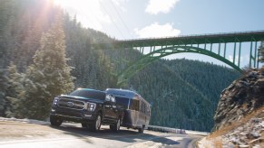 Black Ford F-150 pulls an airstream camper trailer up a steep hill, beneath a bridge, trees visible in the background.