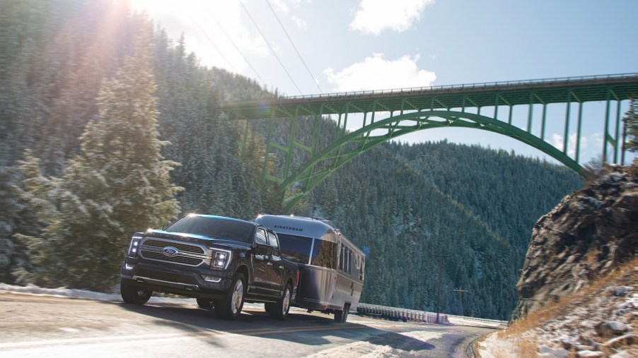 Black Ford F-150 pulls an airstream camper trailer up a steep hill, beneath a bridge, trees visible in the background.