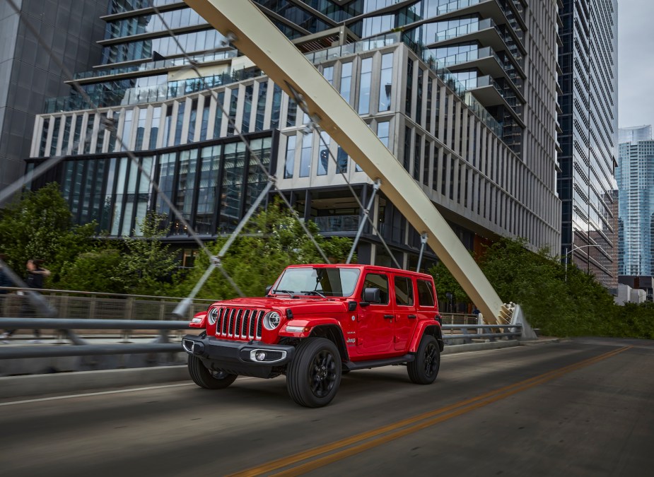 A red hybrid Jeep Wrangler 4xe hybrid SUV driving across a bridge, an American city visible in the background.