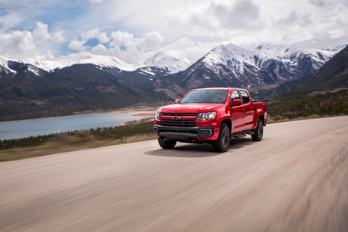 A red 2022 Chevrolet Colorado is driving on wide paved road with a body of water and snow-capped mountains in the background.