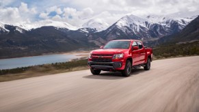 A red 2022 Chevrolet Colorado is driving on wide paved road with a body of water and snow-capped mountains in the background.