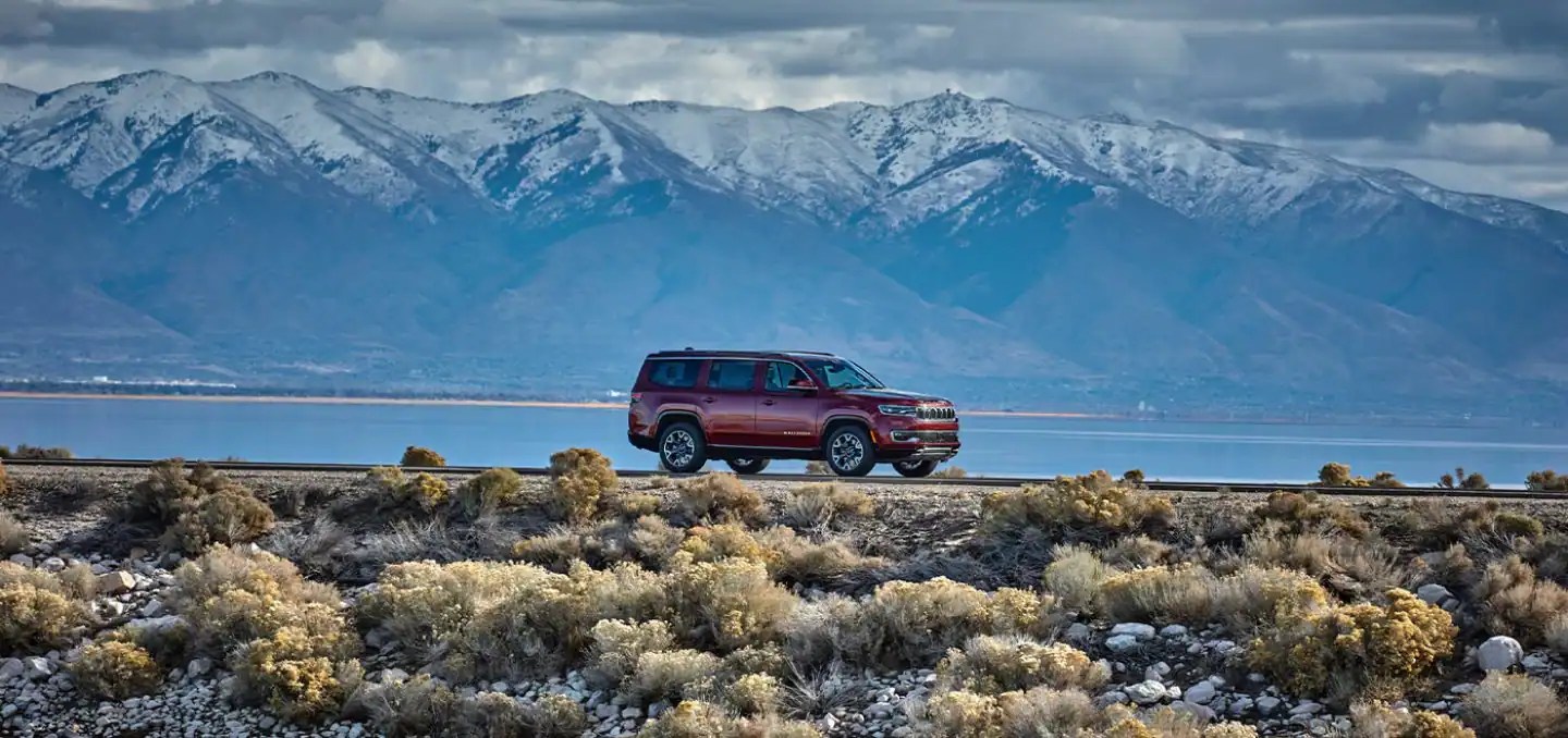 2022 Jeep Grand Wagoneer parked in the midground of the frame in desert terrain. with snow-capped mountains behind it.