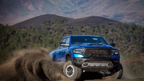 A blue Ram TRX truck skids across an off-road trail, mountains and a cloud of dust visible in the background.