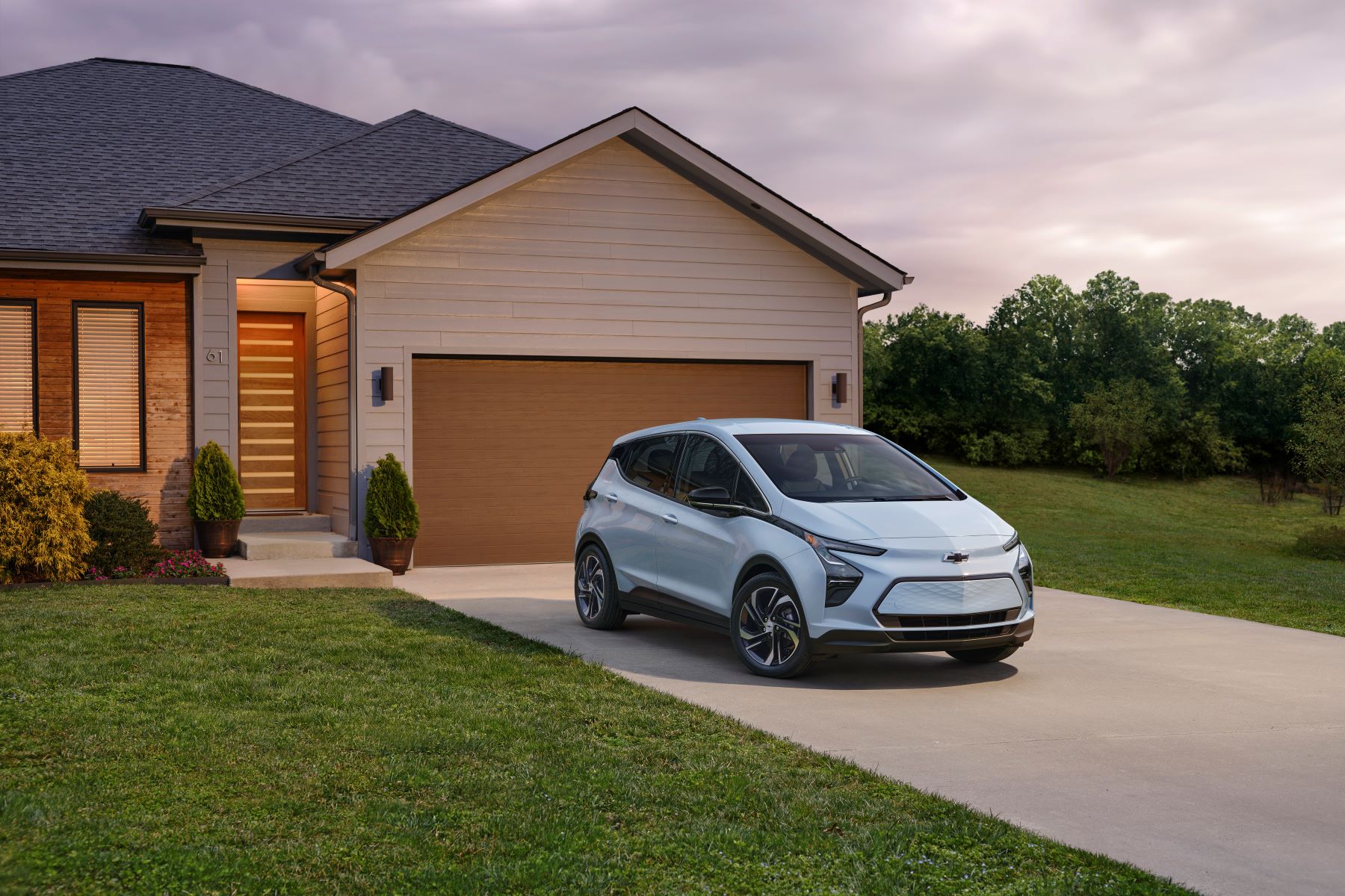 A light blue 2023 Chevy Bolt EV parked outside a home's garage near a cleaning cut grass lawn