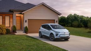 A light blue 2023 Chevy Bolt EV parked outside a home's garage near a cleaning cut grass lawn