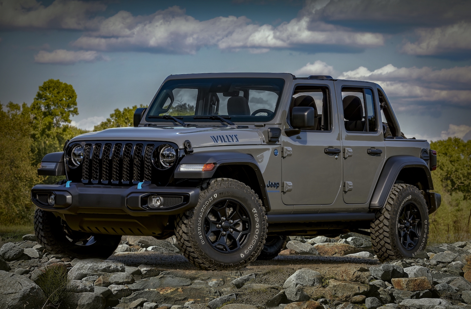 A gray Jeep Wrangler "Willys" edition hybrid 4xe parked on a pile of rocks for a promo photo, trees visible in the background.