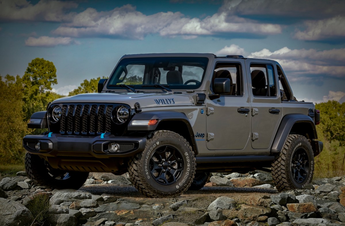 A gray Jeep Wrangler "Willys" edition hybrid 4xe parked on a pile of rocks for a promo photo, trees visible in the background.