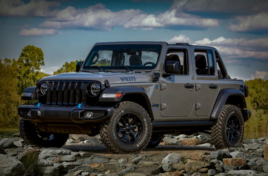 A gray Jeep Wrangler "willy's" 4xe hybrid edition parked on a rock pile for a promotional photo, trees visible in the background.