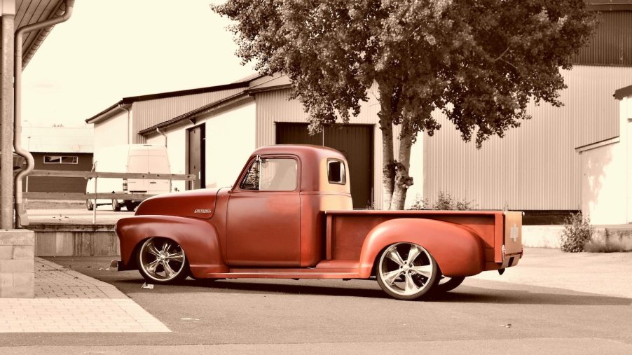 A classic red Chevrolet stepside pickup truck parked in front of a tree, in a parking lot.
