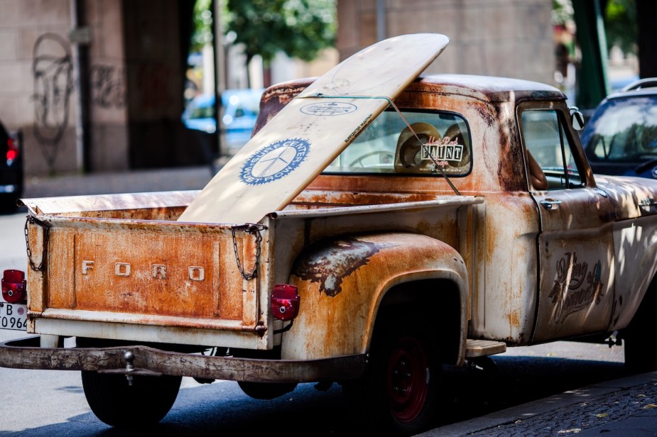 A rusty Ford stepside pickup truck parked on a city street, a surfboard fixed in its bed.