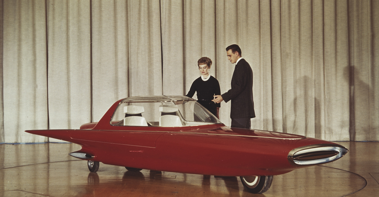A couple stands next to the 1961 Ford Gyron concept car on stage at the Detroit auto show, a white curtain visible behind them. 