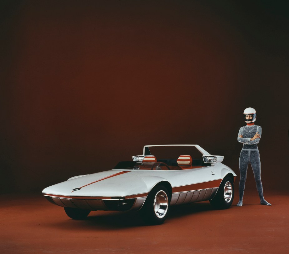 A woman in a helmet stands next to a 1969 Italian concept car with no roof on the stage of the Turin auto show, a black background behind her.