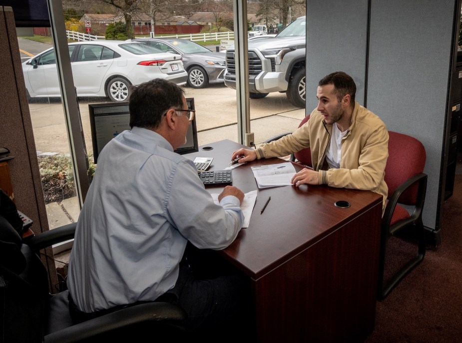 A dealership salesman and a customer talk over his desk about a potential EV or ICE purchase, a lot of cars visible through the window.