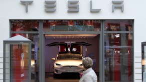 A photo of a woman walking past a corporate-owned Tesla EV showroom, an SUV visible in its window.