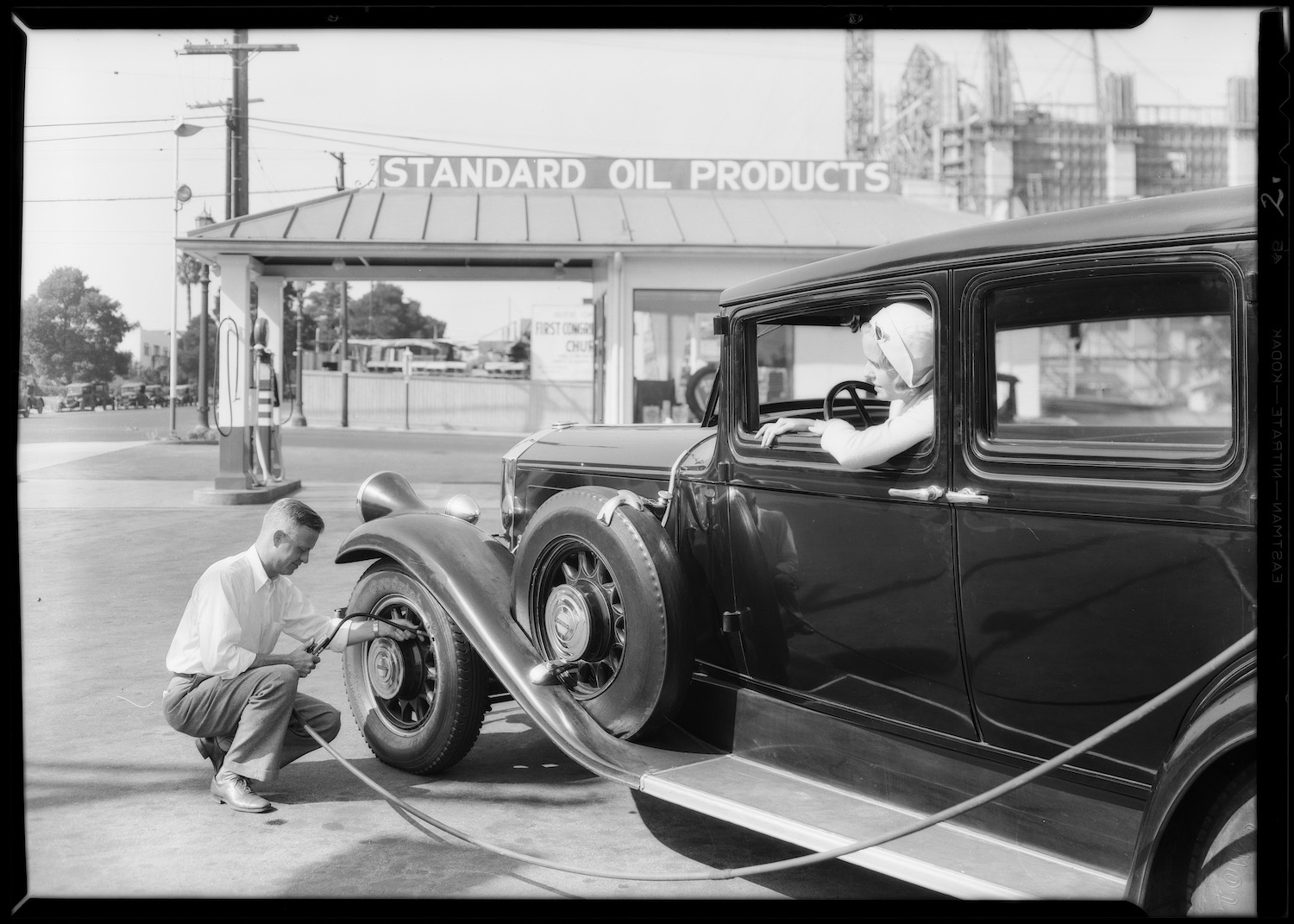 A man crouches by the front tire of a classic car to air up its tires with a hose running from a gas station.
