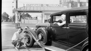 A man crouches by the front tire of a classic car to air up its tires with a hose running from a gas station.