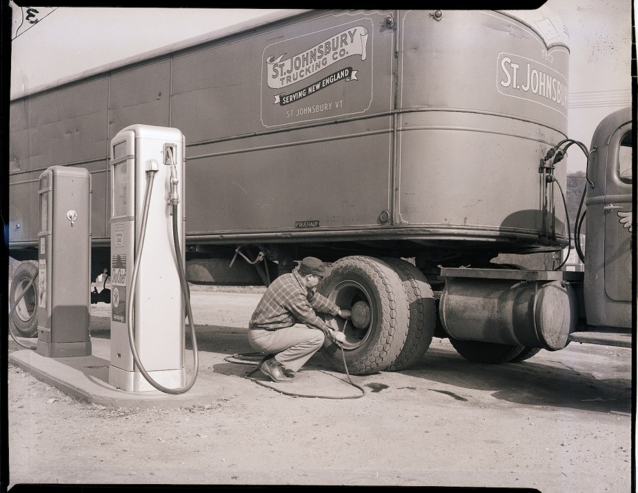 A truck driver squats by his tire to fill it up with air from a gas station's compressor.