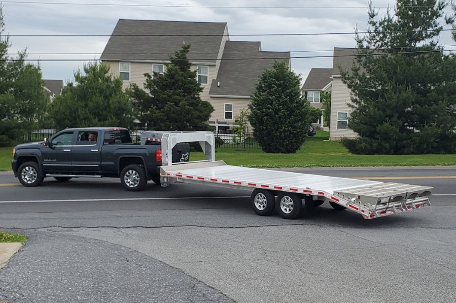 Dark gray heavy-duty GMC pickup truck pulling a flatbed gooseneck trailer down a driveway and onto the street, houses visible in the background.