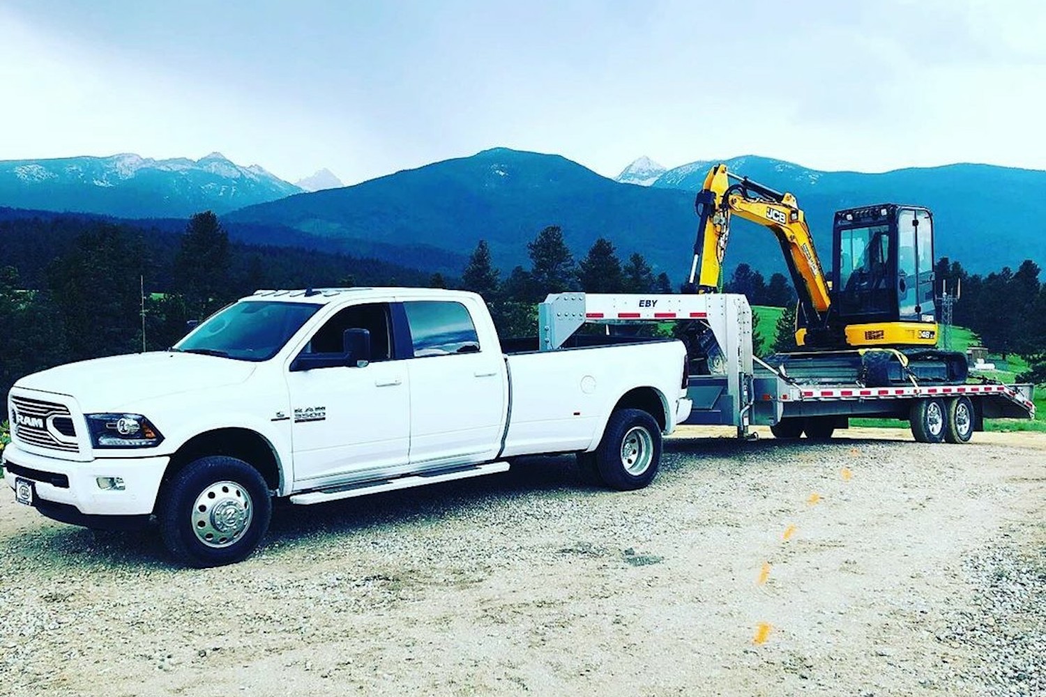 White Ram 3500 heavy-duty double pickup truck pulling an excavator on a flatbed gooseneck trailer, with mountains visible in the background.