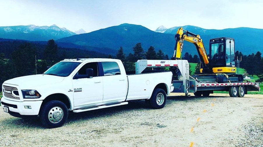 White, heavy-duty Ram 3500 dually truck pulling an excavator on a flatbed gooseneck trailer, mountains visible in the background.