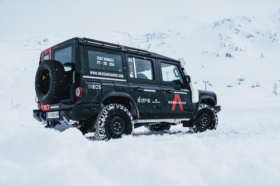 An Ineos Grenadier 4x4 SUV equipped with a snorkel parked off-road on the side of a snowy mountain, with a chairlift visible in the background.