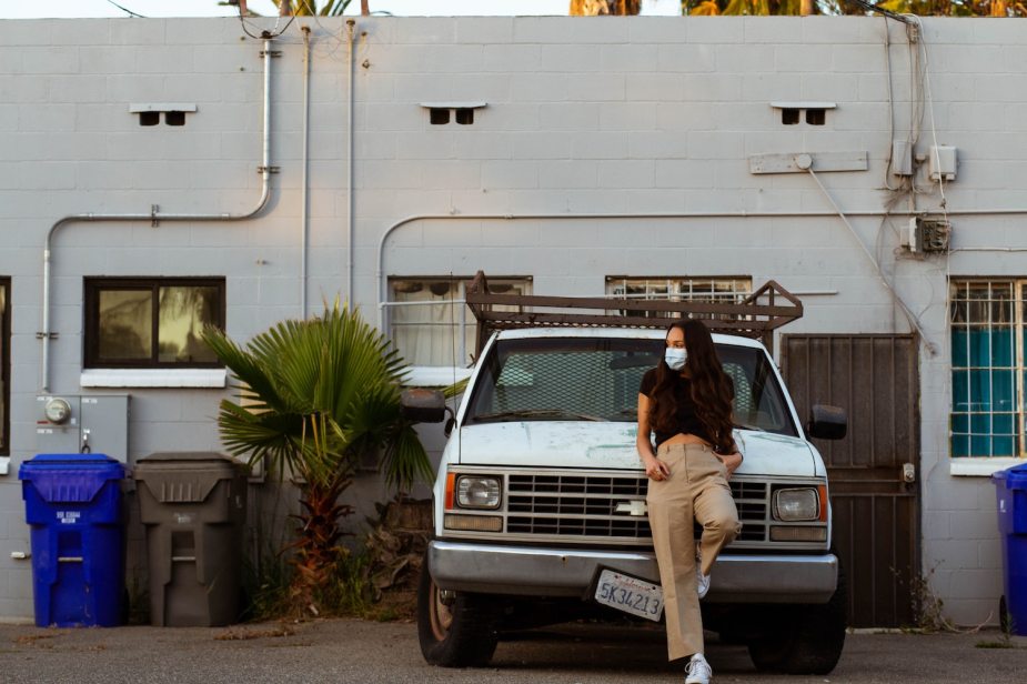 A woman leans against the grille of a a white OBS Chevy work truck which is parked in front of a warehouse, plants and recycling bins visible nearby.