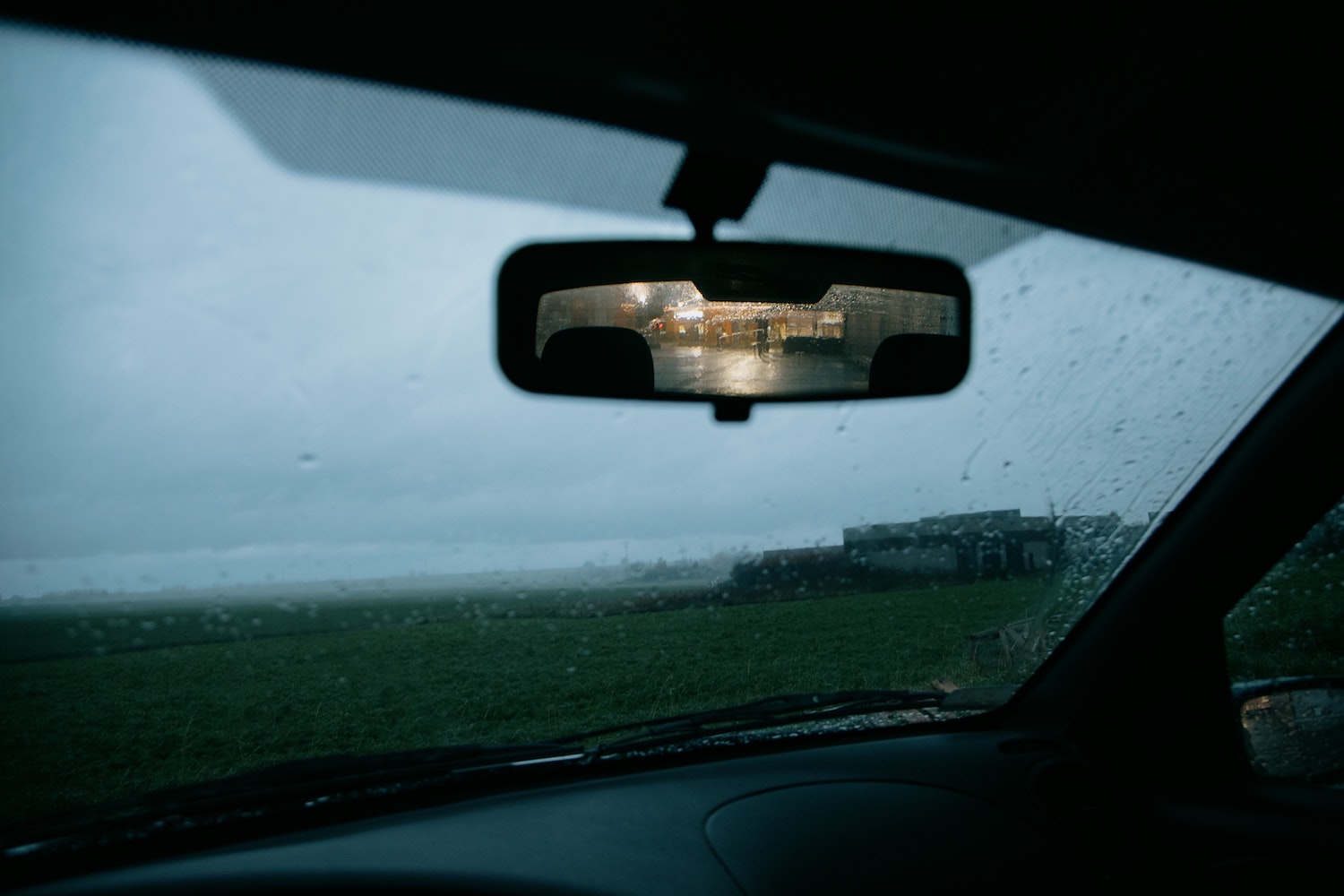 Traffic visible in a car's rearview mirror, a rain-covered windshield behind it.