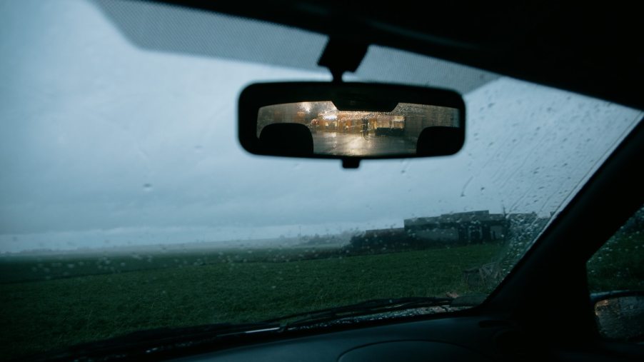 Traffic visible in a car's rearview mirror, a rain-covered windshield behind it.