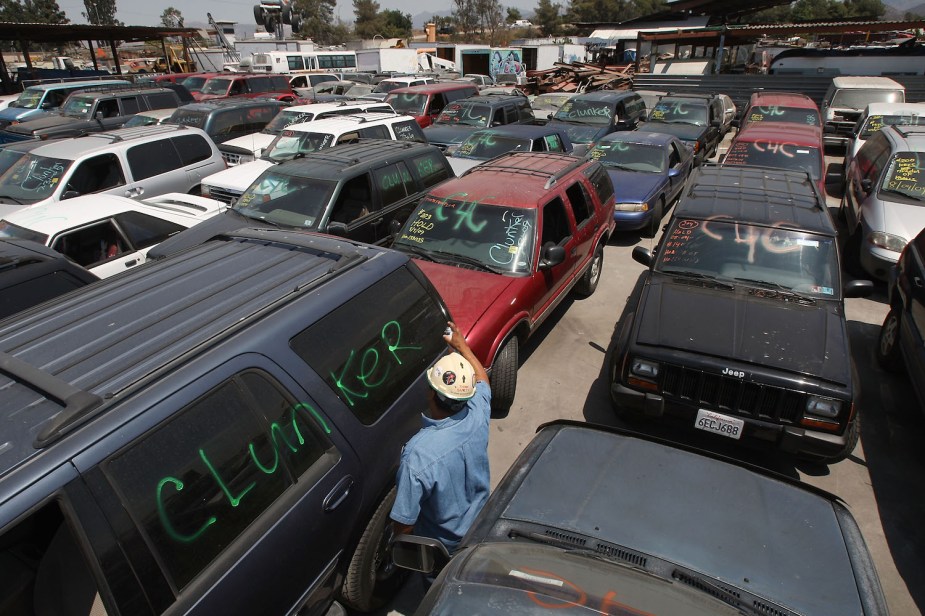 Rows of old cars to be scrapped, parked in front of a junkyard. A man in the foreground spray paints "Clunker" on a window.