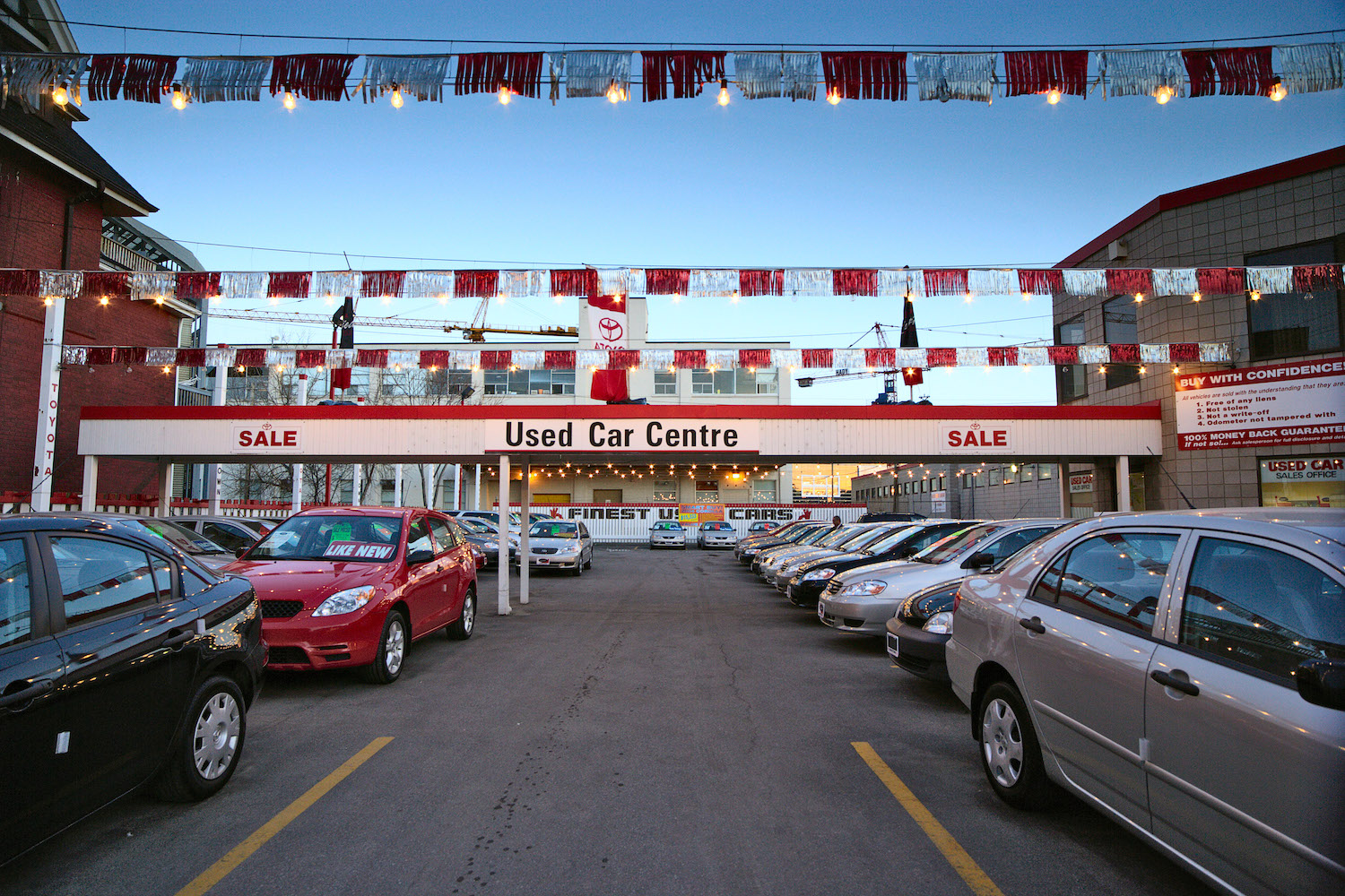 Rows of used cars parked in the lot outside a dealership with red and white banners and a Toyota sign visible in the background.