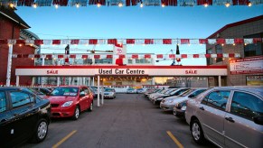 Rows of used cars parked in the lot outside a dealership with red and white banners and a Toyota sign visible in the background.