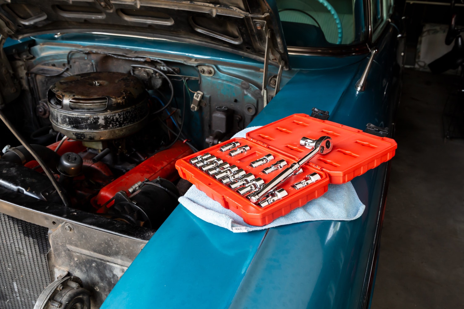 An orange box of sockets and a socket wrench, set on the blue fender of a classic car, its V8 engine visible in the background.