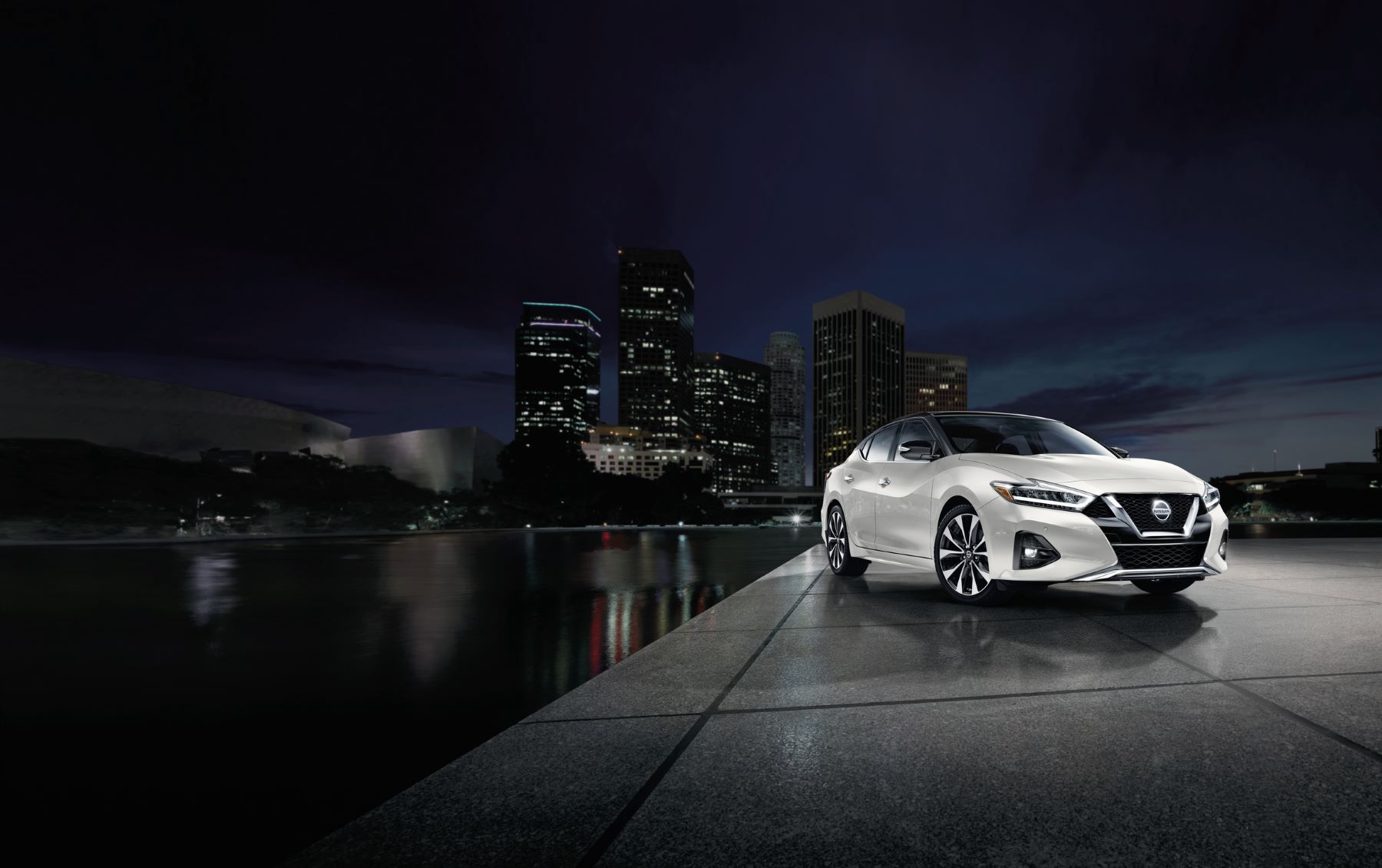 A white 2023 Nissan Maxima midsize sedan parked on a marble plaza near water with a city in the background at night