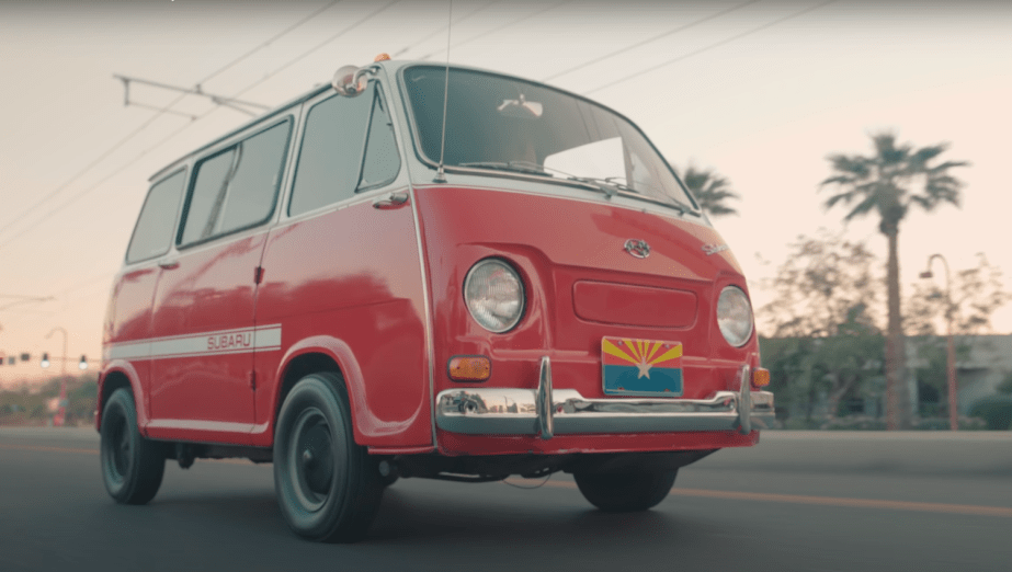 A red Subaru Sambar microbus drives down a road in Los Angeles, palm trees visible in the background.