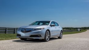 A 2015 Acura TLX entry-level luxury sedan model parked on a gravel road near metal fencing on a farm. The TLX is one of the most reliable Acura models
