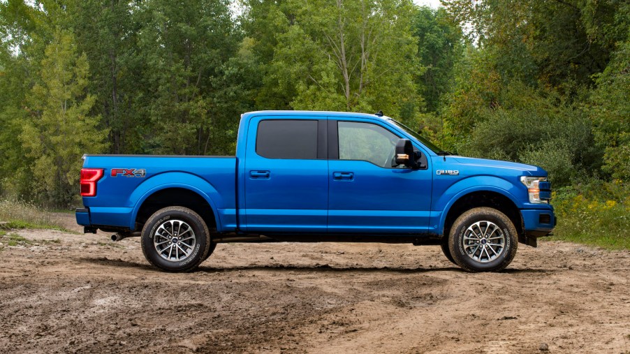 Blue Ford F-150 full-size pickup truck parked on a dirt parking lot, trees visible in the background.